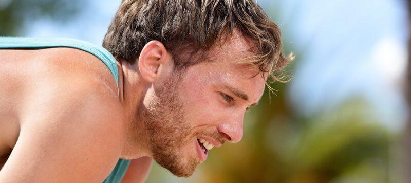 Tired exhausted man runner sweating after cardio workout. Running male adult taking a break and breaking a sweat after a run under the sun. Fitness athlete breathing heavily from heat exhaustion.